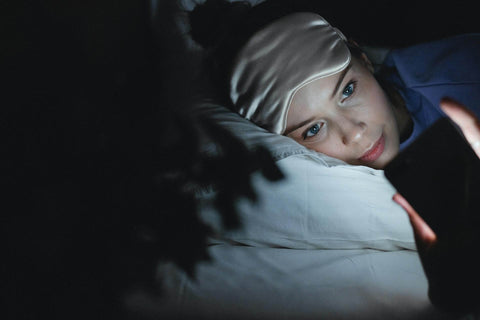 Vrouw met een slaapmasker die 's nachts naar haar telefoon kijkt, wat de effecten van blauw licht illustreert.